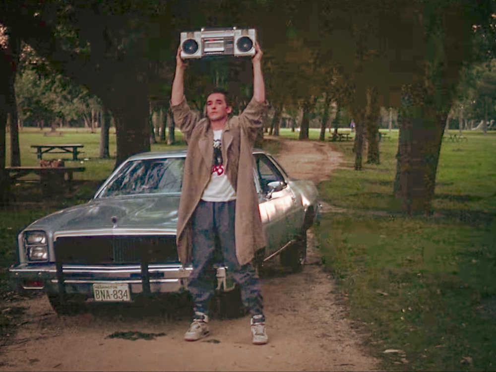 Man stands in front of a car holding a boombox above his head