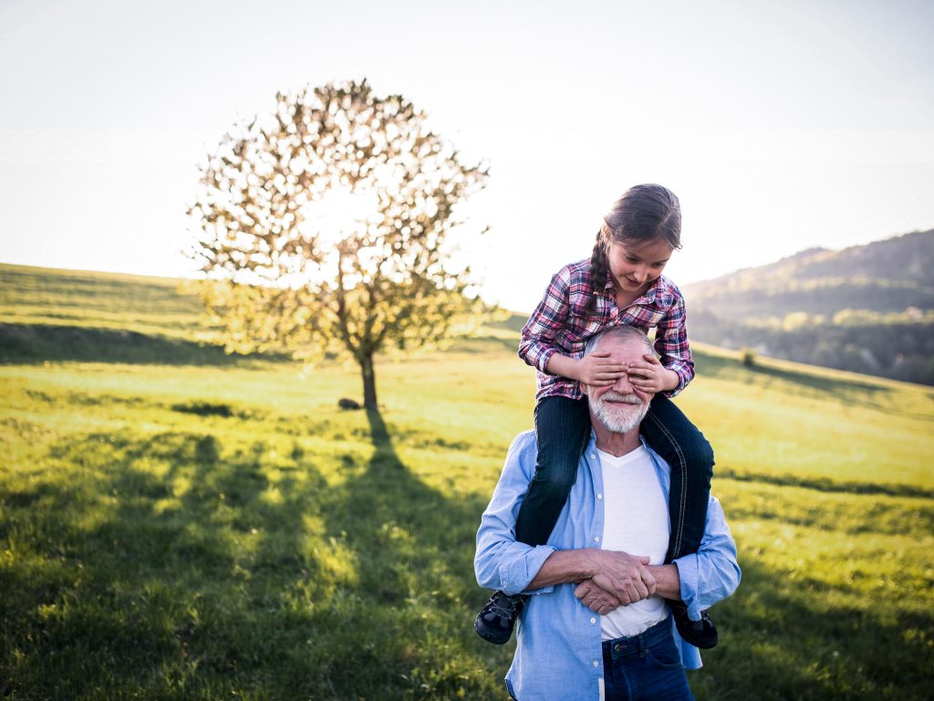 Grandfather carries granddaughter on shoulders in front of a tree