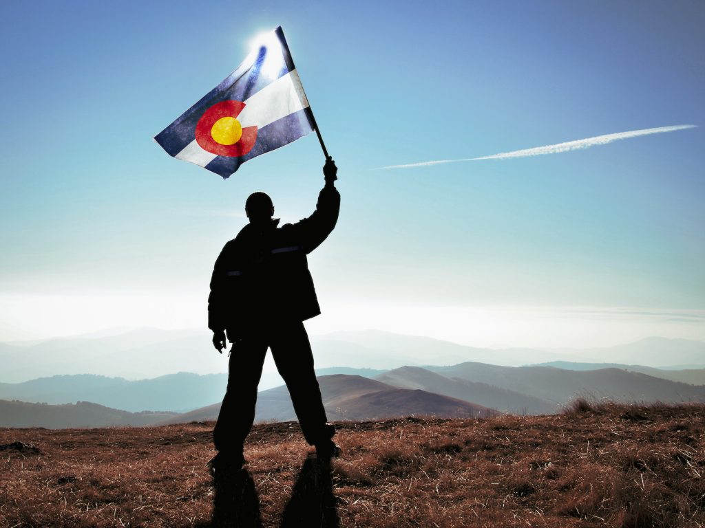 man stands on a plateau with Colorado flag raised
