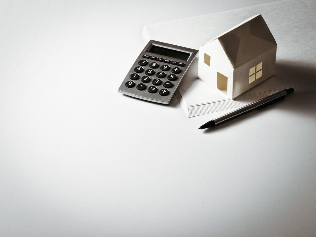 A miniature house sits on a table next to a calculator, pen and pad of paper