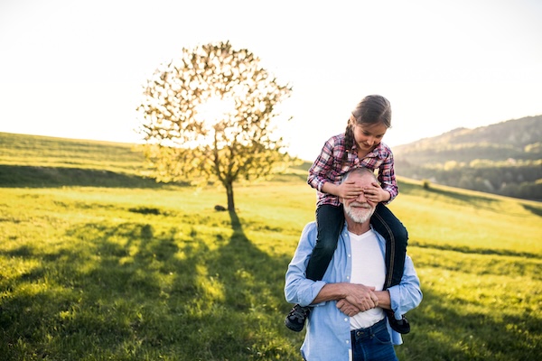 grandfather holds granddaughter on his shoulders in front of a tree