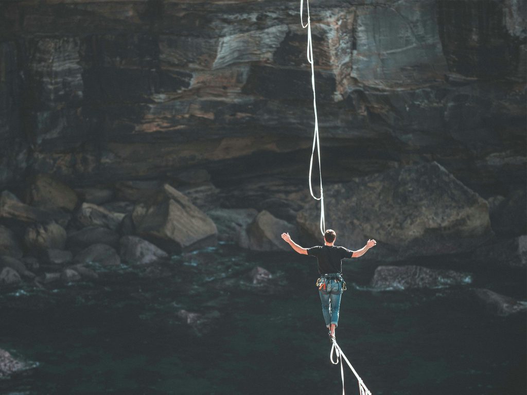 A person is walking on a highline rope above rocks and water, balancing with arms outstretched.